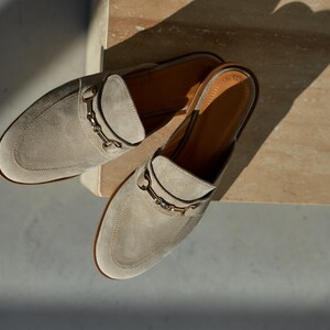 May include: A pair of light gray suede slip-on mules with a gold-colored metal accent on the vamp. The shoes have a rounded toe and a low heel. The interior is a light brown color.