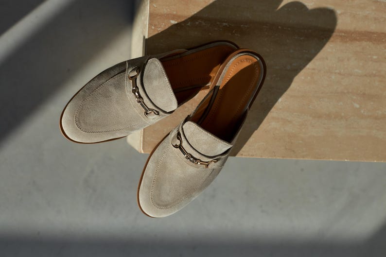 May include: A pair of light gray suede slip-on mules with a gold-colored metal accent on the vamp. The shoes have a rounded toe and a low heel. The interior is a light brown color.