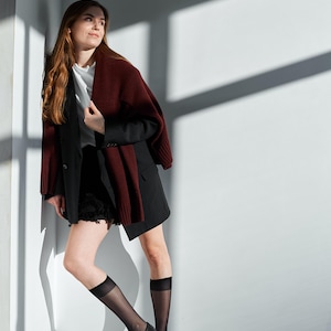 May include: A woman wearing a black blazer, black shorts, and black shoes. A burgundy sweater is draped over the shoulders. She is also wearing sheer black knee-high socks. The background is a white wall with shadows.