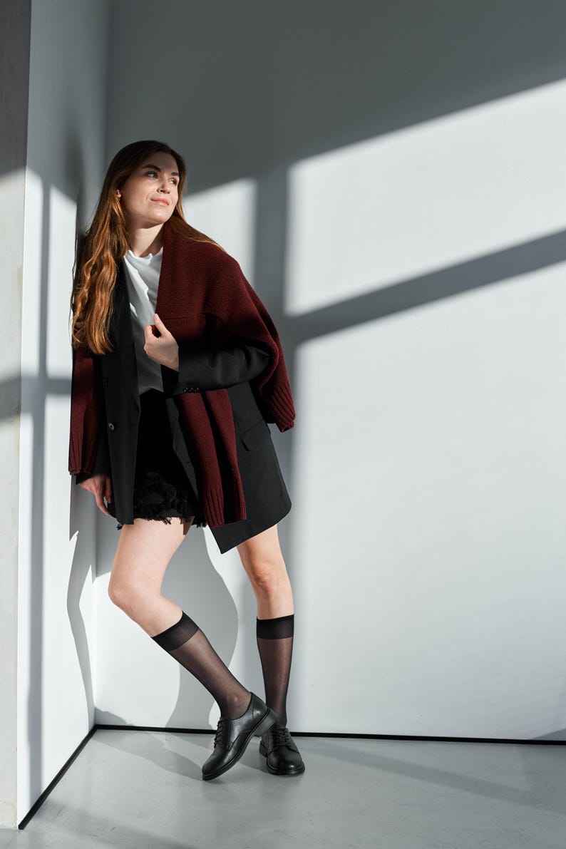 May include: A woman wearing a black blazer, black shorts, and black shoes. A burgundy sweater is draped over the shoulders. She is also wearing sheer black knee-high socks. The background is a white wall with shadows.