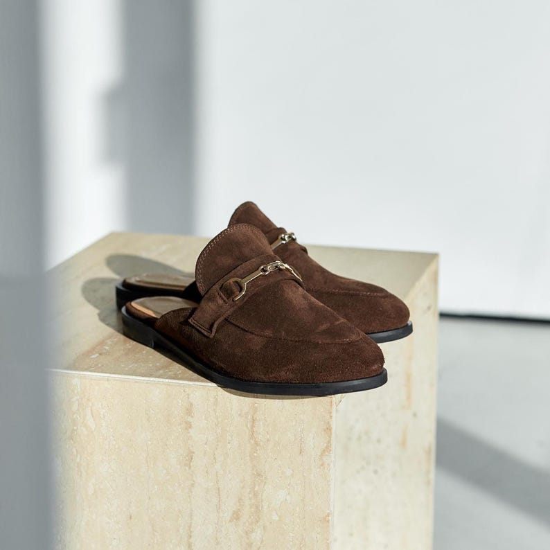 May include: A pair of brown suede slip-on shoes with a gold-colored metal buckle detail. The shoes have a rounded toe and a flat sole. The shoes are displayed on a light-colored surface.