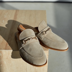 May include: A pair of beige suede slip-on loafers with gold-colored metal accents. The shoes have a rounded toe and a low heel. The loafers are displayed on a light-colored, textured surface.