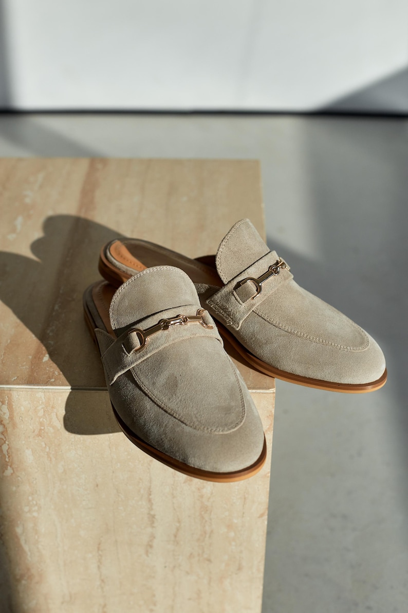 May include: A pair of beige suede slip-on loafers with gold-colored metal accents. The shoes have a rounded toe and a low heel. The loafers are displayed on a light-colored, textured surface.