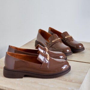 May include: Brown patent leather loafers with a gold chain detail. The shoes have a rounded toe and a low, chunky heel. The loafers are displayed on a light-colored surface.