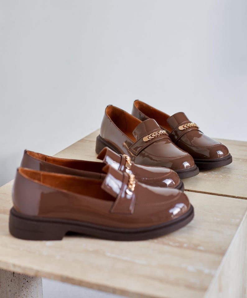 May include: Brown patent leather loafers with a gold chain detail. The shoes have a rounded toe and a low, chunky heel. The loafers are displayed on a light-colored surface.