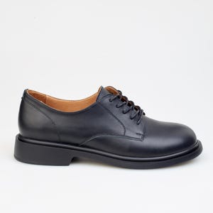 May include: A single black leather oxford shoe with a rounded toe and a thick sole. The shoe has a lace-up closure and a low heel. The interior lining is a light brown color. The shoe is set against a white background.
