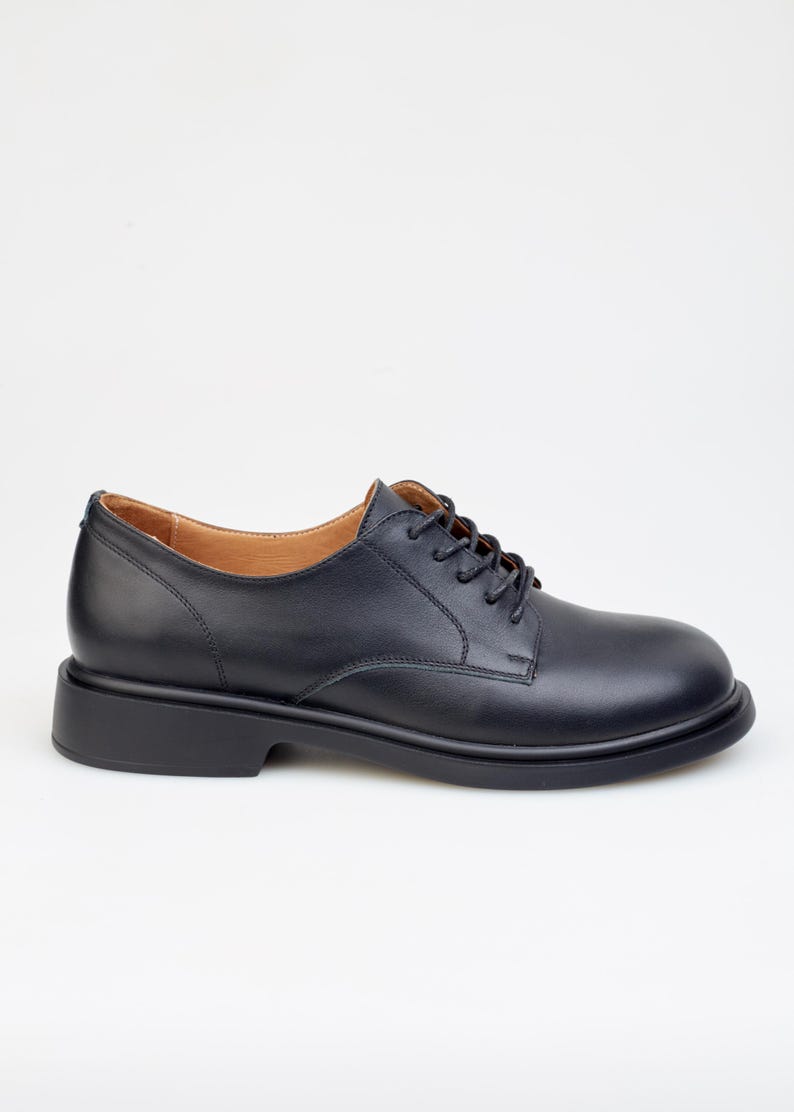 May include: A single black leather oxford shoe with a rounded toe and a thick sole. The shoe has a lace-up closure and a low heel. The interior lining is a light brown color. The shoe is set against a white background.