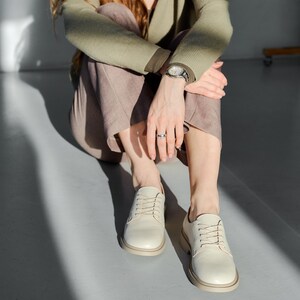 May include: Cream-colored leather shoes with a rounded toe and a thick sole. The shoes are laced up and appear to be new. The shoes are on a light gray surface.