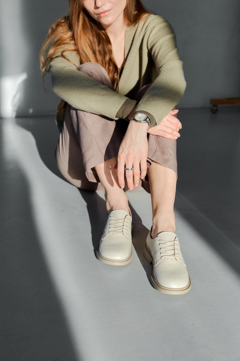 May include: Cream-colored leather shoes with a rounded toe and a thick sole. The shoes are laced up and appear to be new. The shoes are on a light gray surface.