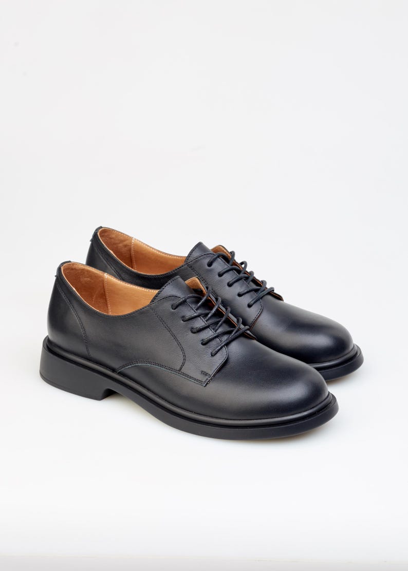May include: A pair of black leather Oxford shoes with a rounded toe and a thick sole. The shoes have black laces and a light brown interior. The shoes are displayed on a white background.
