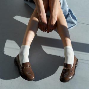 May include: A pair of brown patent leather loafers with gold chain detailing. The shoes are worn with white socks. The image is taken from a low angle, with the person wearing the shoes sitting on a gray surface.