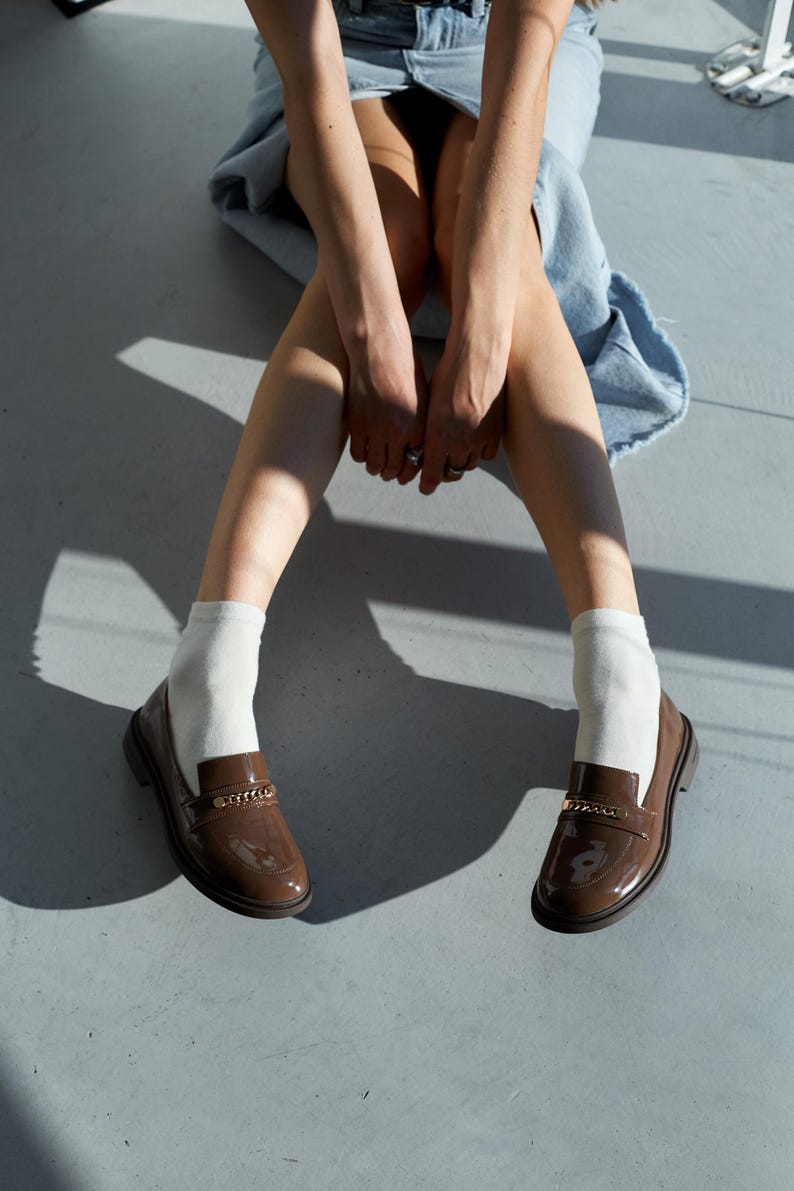 May include: A pair of brown patent leather loafers with gold chain detailing. The shoes are worn with white socks. The image is taken from a low angle, with the person wearing the shoes sitting on a gray surface.