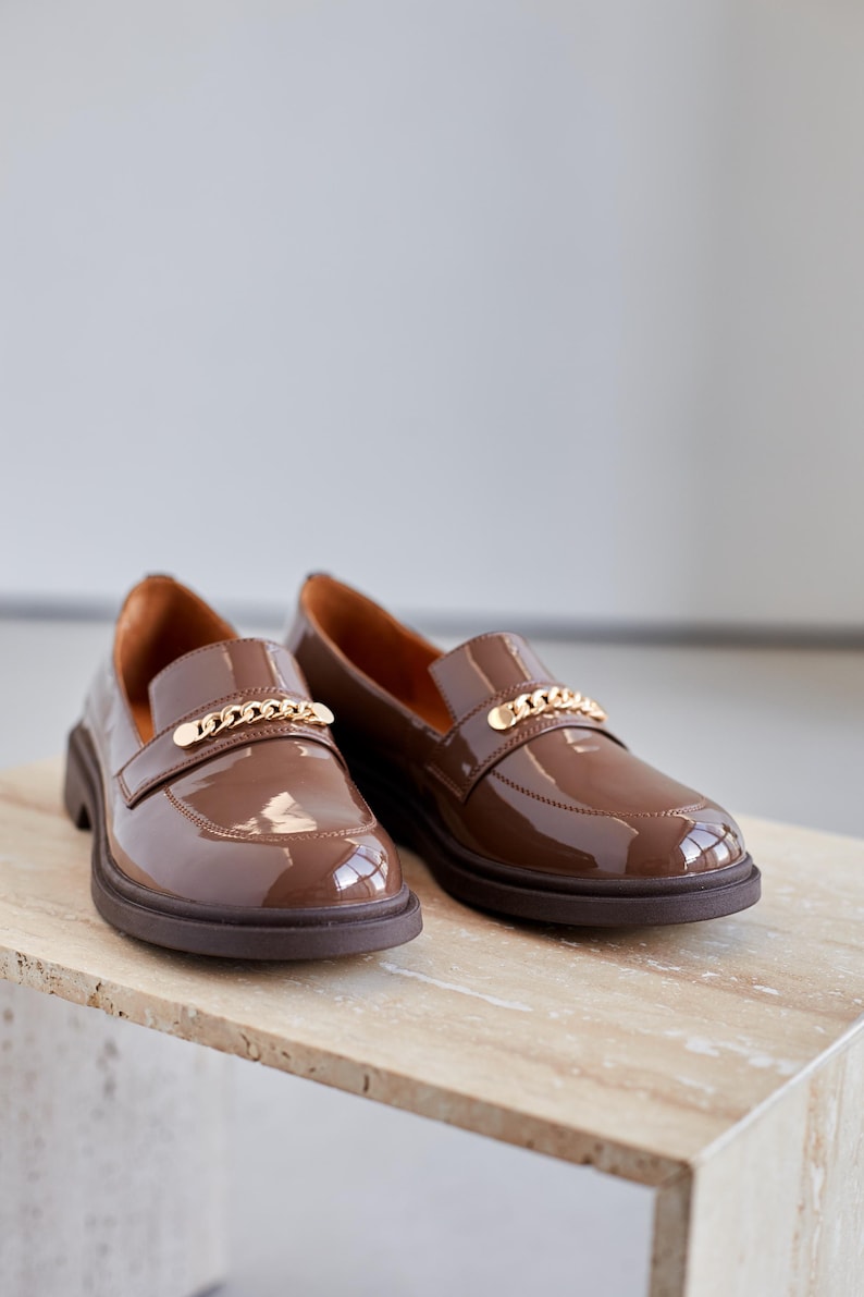 Women’s brown patent leather loafers with a glossy finish and chunky sole. Designed with a gold chain detail across the upper for a modern, stylish look. Slip-on style makes them versatile and comfortable for everyday wear, office outfits, or casual chic streetwear. Durable sole provides support while the polished surface adds elegance, making these loafers a perfect blend of sophistication and practicality for fashion-forward women.