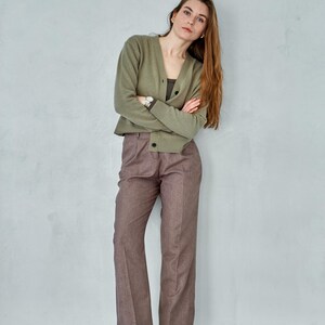 May include: A woman wearing a sage green cardigan, a dark green top, and brown trousers. She is also wearing white shoes. The outfit is simple and elegant, suitable for a casual or semi-formal setting.