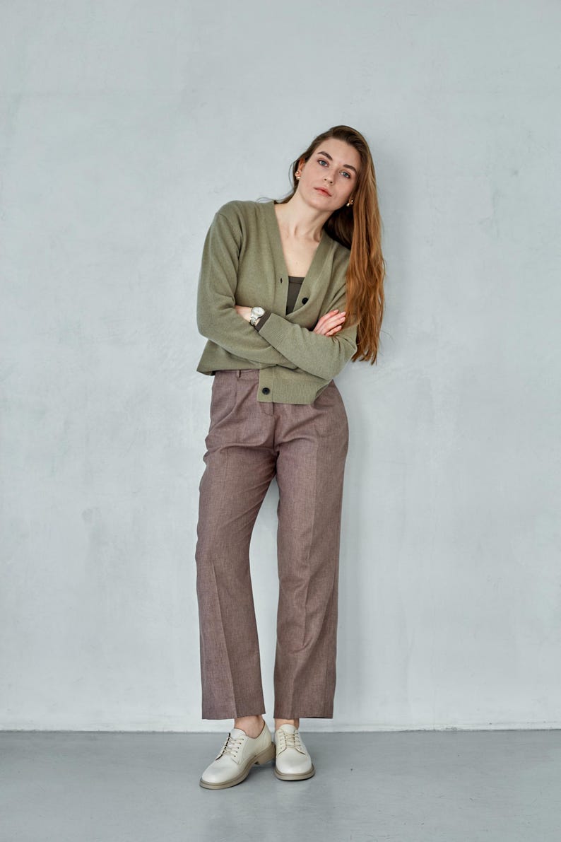 May include: A woman wearing a sage green cardigan, a dark green top, and brown trousers. She is also wearing white shoes. The outfit is simple and elegant, suitable for a casual or semi-formal setting.