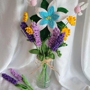 May include: A handmade crochet bouquet featuring a large white and blue flower, yellow and purple flowers, and green leaves, arranged in a clear glass jar tied with twine. Two lavender stems are placed in front of the jar. The bouquet includes two pink and white fairy-like accents.