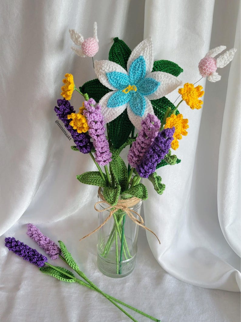 May include: A handmade crochet bouquet featuring a large white and blue flower, yellow and purple flowers, and green leaves, arranged in a clear glass jar tied with twine. Two lavender stems are placed in front of the jar. The bouquet includes two pink and white fairy-like accents.