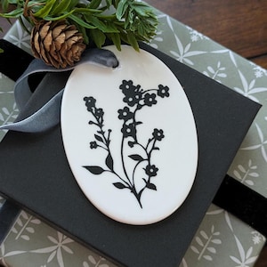 May include: A white oval ornament with a black floral design, tied with a gray ribbon and placed on a black gift box. The gift box is on top of a patterned gift wrap. A pine cone and evergreen sprig add a natural touch.