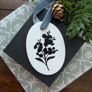May include: An oval white ornament with a black floral design, hanging from a gray ribbon. The ornament rests on a black box, atop a gift box wrapped in gray and white patterned paper. A pinecone and greenery add a natural touch.