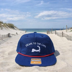May include: A navy blue cap with a red brim and the text "ESCAPE TO THE CAPE" in white. The cap features a white outline of a peninsula. The cap is sitting on a sandy beach.