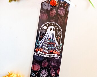 May include: A black bookmark with a ghost holding a stack of books. The bookmark has a fall theme with pumpkins, leaves, and snowflakes. The text on the bookmark reads "Haunted by my TBR".