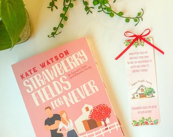 May include: A pink paperback book titled "Strawberry Fields for Never" by Kate Watson. The book cover features a cartoon illustration of a couple standing in front of a red barn and a fence. A red bookmark with a quote and a farm illustration is tucked into the book.