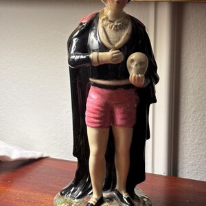May include: A porcelain figurine of a figure wearing a black cap, a black cape, and pink shorts, holding a skull in one hand. The figure stands on a base with the text "Alas poor Yorick!" inscribed on the front, featuring painted details.