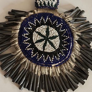 May include: A circular medallion featuring intricate beadwork in blue, white, and black, arranged in a starburst pattern. The piece is bordered by numerous metallic, cone-shaped fringe elements attached to a light-colored leather backing.