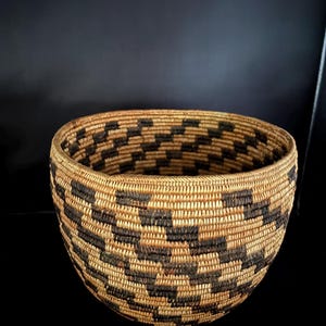 May include: A round, hand-woven basket featuring a repeating geometric pattern in dark brown and natural tan fibers. The basket has a wide, open top and a deep, bowl-shaped body constructed with tightly coiled, horizontal rows of woven plant material.