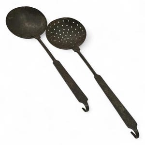 May include: Two antique, dark metal kitchen utensils. One is a solid, round ladle, and the other is a slotted skimmer. Both have long handles with a hook at the end for hanging. The utensils are likely made of iron.