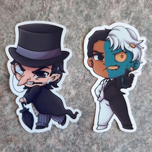 May include: Two cartoon stickers of DC Comics villains. One shows The Penguin in a black top hat and coat, holding an umbrella. The other depicts Two-Face, with one side of his face orange and the other green.