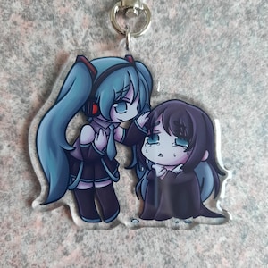 May include: A clear acrylic keychain featuring two anime-style characters. One character has long blue hair and is wearing a black outfit. The other character has dark hair and is crying. The keychain has a silver clasp.