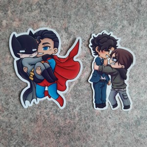 May include: Two cartoon stickers featuring comic book characters. One shows Superman carrying Batman, the other depicts two men in a heated embrace. The stickers have a white border and are set against a mottled gray background.