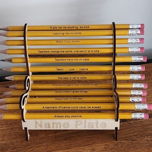 Peut inclure: Un présentoir en bois avec des crayons jaunes disposés en gradins. Chaque crayon, avec une gomme, porte des citations inspirantes. Le nom du présentoir est visible en bas.