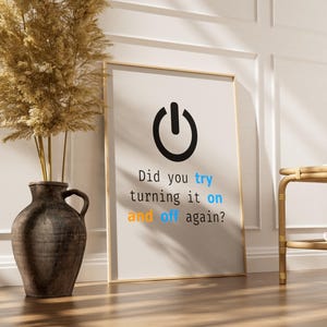 May include: A framed print with a black power button icon and the text "Did you try turning it on and off again?" in a modern font. The print is leaning against a wall, next to a vase with dried plants.