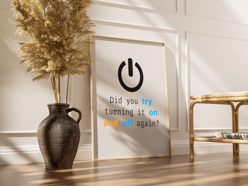May include: A framed print with a black power button icon and the text "Did you try turning it on and off again?" in a modern font. The print is leaning against a wall, next to a vase with dried plants.