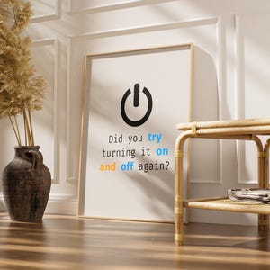 May include: A framed print with a black power button icon and the text "Did you try turning it on and off again?" in a modern font. The print is leaning against a white wall. A decorative vase and a small table are also in the image.