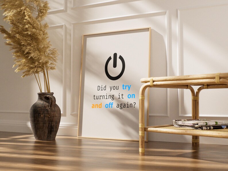 May include: A framed print with a black power button icon and the text "Did you try turning it on and off again?" in a modern font. The print is leaning against a white wall. A decorative vase and a small table are also in the image.