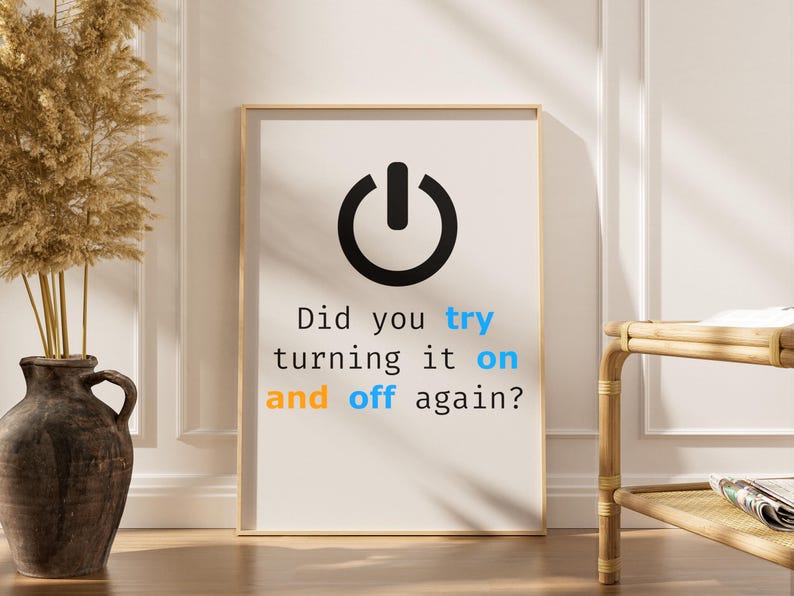 May include: A framed print with a black power button icon and the text "Did you try turning it on and off again?" in black, blue, and orange. The print is in a light wood frame.