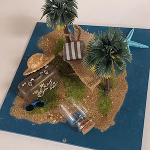 Könnte beinhalten: Miniatur-Insel-Diorama mit Sand, Palmen und Reisezubehör. Enthält einen braunen Koffer mit Text, einen Strohhut, eine Sonnenbrille, eine gestreifte Tasche, eine Glasflasche und einen blauen Seestern auf blauem Grund.
