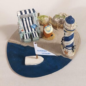 Könnte beinhalten: Eine Miniatur-Strandszene mit einem blau-weiß gestreiften Strandkorb, einem Leuchtturm und einem kleinen Segelboot mit dem Text "Schönen Urlaubl". Die Szene umfasst ein kleines Haus und Felsen auf einer sandigen Basis.