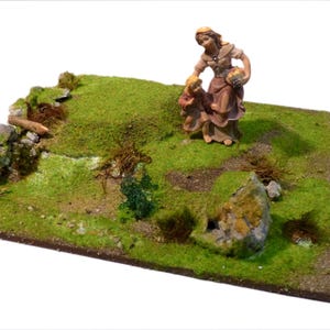 Könnte beinhalten: Ein Miniatur-Diorama mit einem grasbewachsenen Hügel mit einer Steinmauer und einer kleinen Figur einer Frau und eines Kindes. Die Szene ist detailliert mit grünem Gras, kleinen Felsen und einer braunen Basis, die eine rustikale, ländliche Umgebung schafft.
