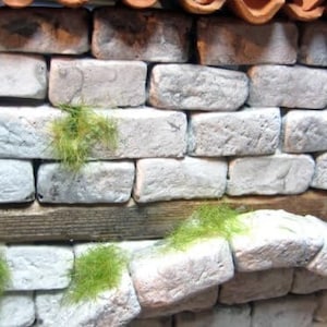 Könnte beinhalten: Eine Miniatur-Steinmauer mit einem Holzbalken und grünem Laub. Die Ziegel sind hellgrau und haben eine raue Textur. Die Mauer wird von terrakottafarbenen Dachziegeln gekrönt.