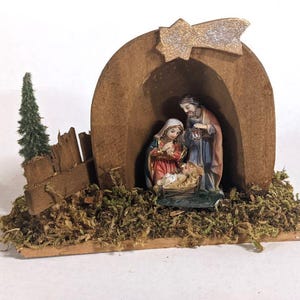 Könnte beinhalten: Eine Miniatur-Weihnachtskrippe mit Maria, Josef und dem Jesuskind in einer Krippe, untergebracht in einem Holzstall. Ein Stern und ein kleiner Nadelbaum sind ebenfalls enthalten. Die Szene befindet sich auf einem mit Moos bedeckten Sockel.