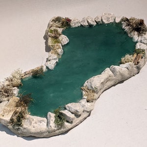 Könnte beinhalten: Miniatur-Teich-Diorama mit türkisfarbener Wasseroberfläche, umgeben von hellen Felsen und Sand. Der Teichrand ist mit kleinen Pflanzen und Moos dekoriert, wodurch eine naturalistische Szene entsteht. Ideal für Modelleisenbahnen oder Miniaturszenen.