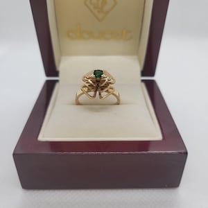 Peut inclure: Une bague en or ornée d'une pierre précieuse verte émeraude, sertie dans un motif ajouré décoratif. La bague est présentée dans un écrin à bijoux marron ouvert, avec la marque "doucet" visible à l'intérieur.