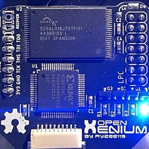 May include: A close-up of a dark blue circuit board with various electronic components. The board features the text "OPEN XENIUM BY RYZEE119" and several chips, including one from Spansion and Xilinx. The board has multiple connection points and a glowing blue LED.