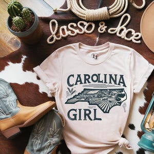 Puede incluir: Una camiseta de color beige claro con un gráfico del estado de Carolina del Norte y el texto "Carolina Girl". El gráfico es en blanco y negro y presenta una granja y campos.