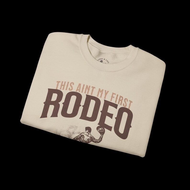 Peut inclure: Un t-shirt beige avec un graphique brun qui dit "This ain't my first rodeo" avec un cowboy chevauchant un taureau.