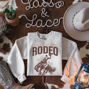 Peut inclure: Un sweat-shirt blanc avec un graphique brun d'un cowboy chevauchant un bronco. Le texte "This ain't my first rodeo" est imprim&eacute; en brun au-dessus du graphique.